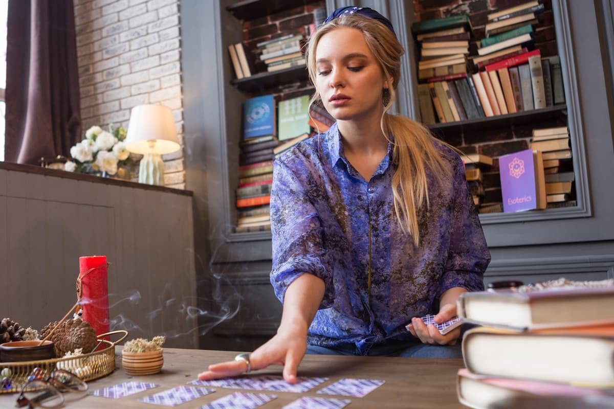 Une jeune femme blonde qui fait un tirage de carte de tarot ou d'oracle sur une table avec des bougies devant une biliothèque